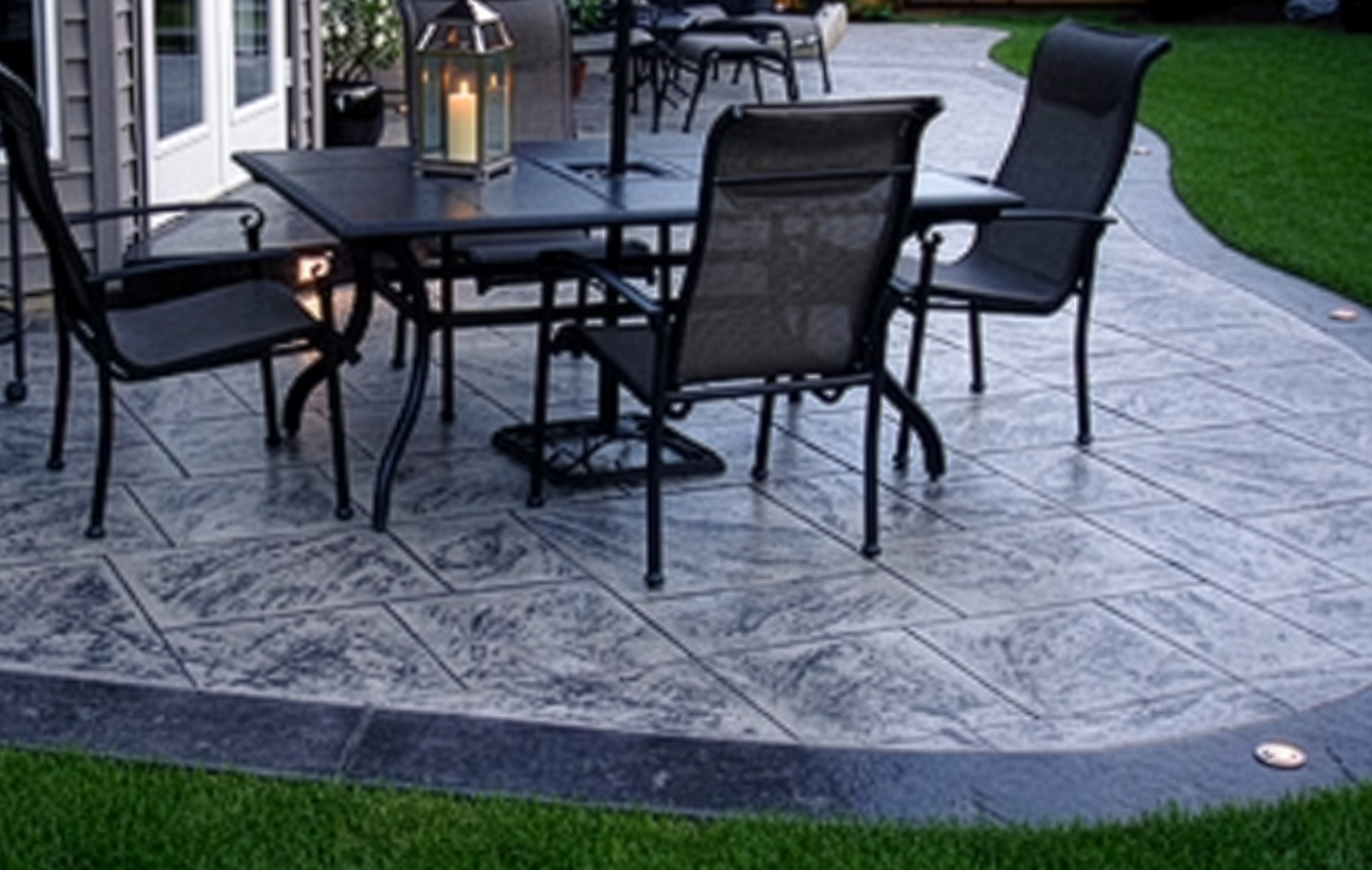 A curved concrete stamp patio with the appearance of being made with light gray flagstones with dark gray streaks. The border of the patio is a dark, charcoal gray. Decorative furniture adorns the patio. 